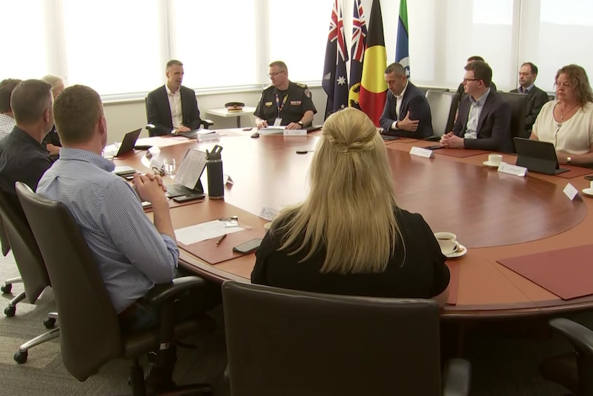 A man speaks at a table surrounded by other people and flags