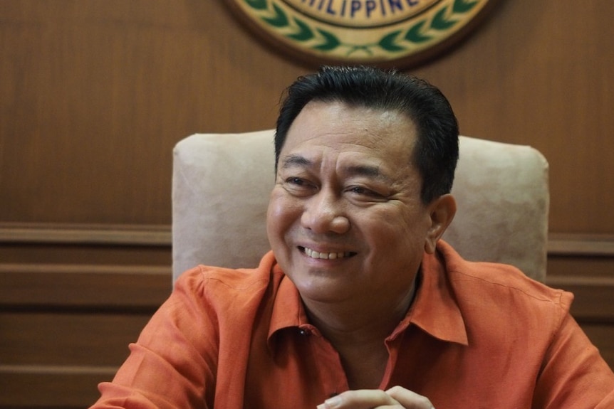 A man laughing wearing an orange shirt in front of the seal for house of representative in the Philippines