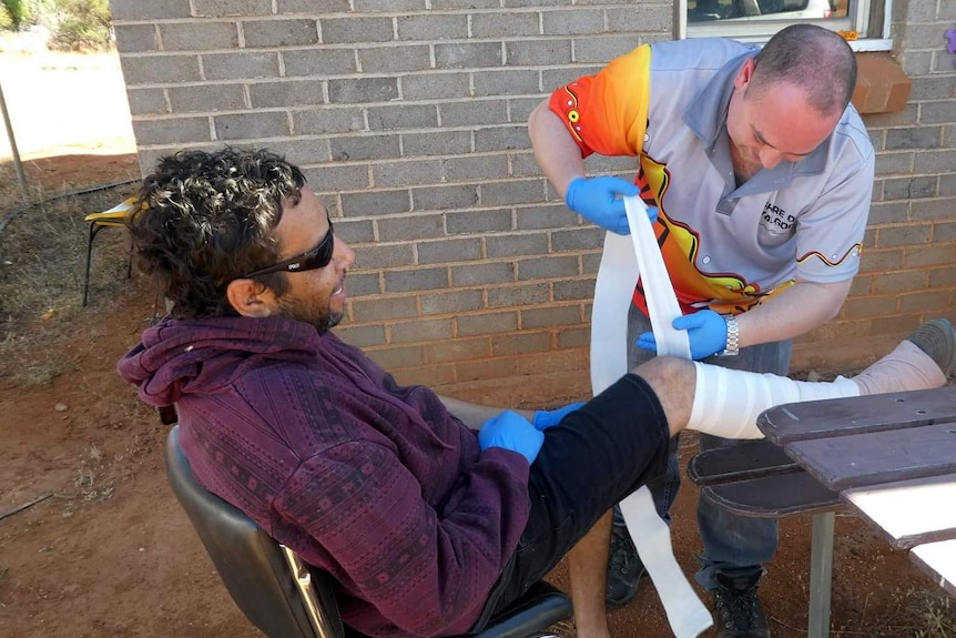 A man applies a leg bandage to another man seated outside.