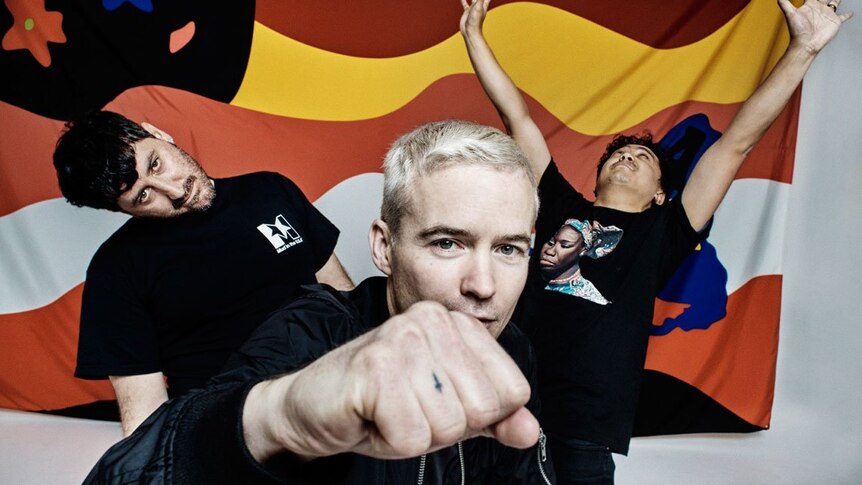 The Avalanches 2016 members in front of colourful waved flag. Robbie pushes his fist forward, James has arms raised