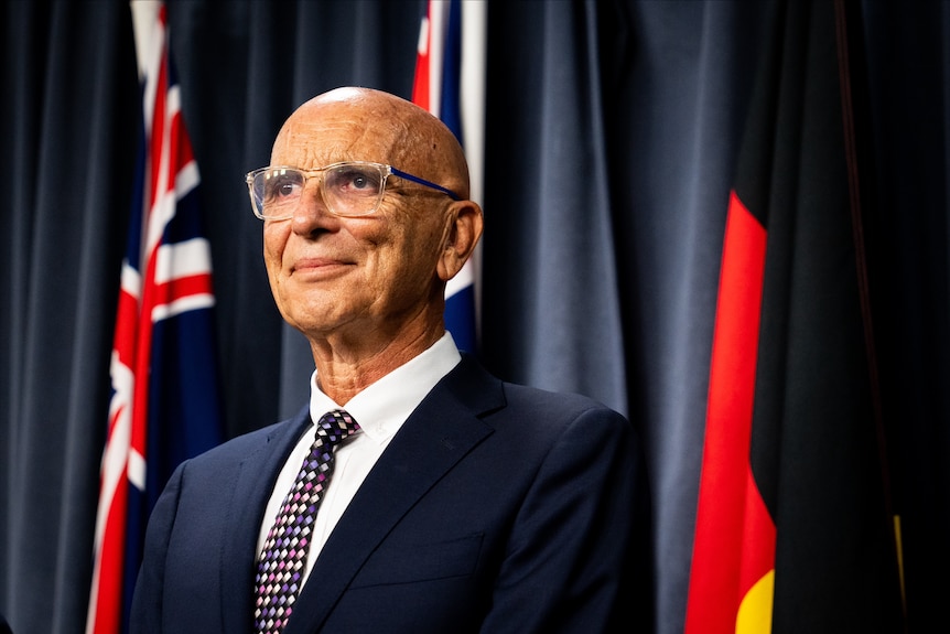 A man wearing glasses smiling while standing in front of flags