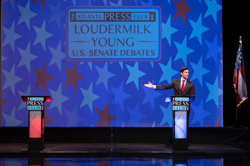 Jon Ossoff stands behind a lectern on a stage gesturing to another empty lectern on his right