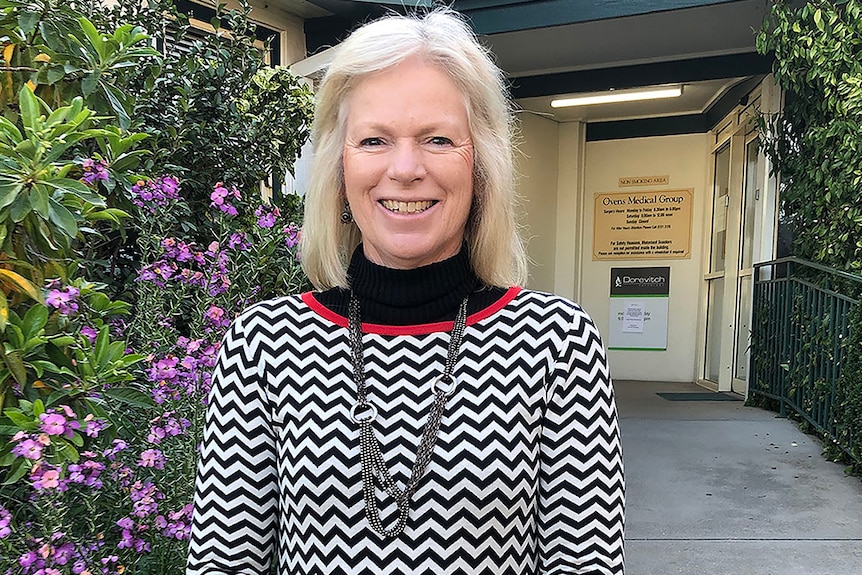 woman with blond hair wearing black and white patterned top stands outside gp clinic