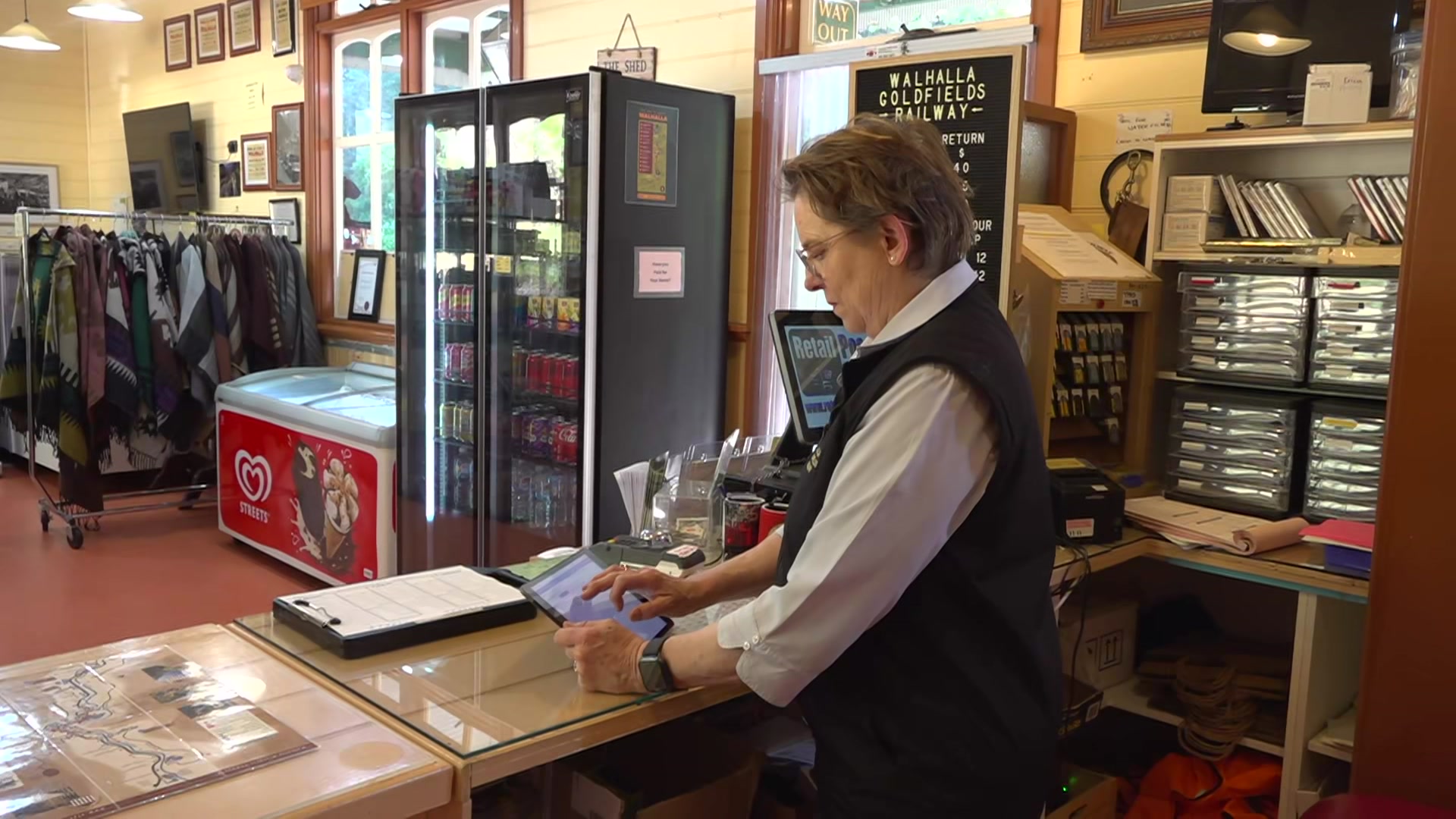 An older lady in a navy uniform looks at an ipad screen behind a shop counter.