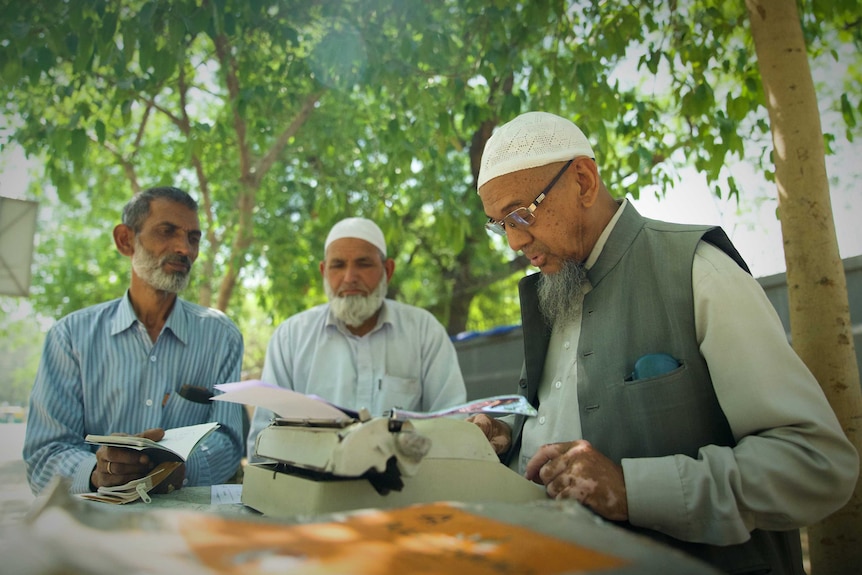 A man stands under a tree on a sunny day typing on a typewriter while two other men look on