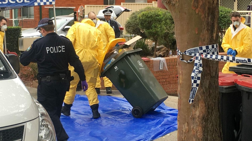 Counter-terrorism police, some dressed in yellow protective suits, outside a Lakemba property. One is dragging a bin away.