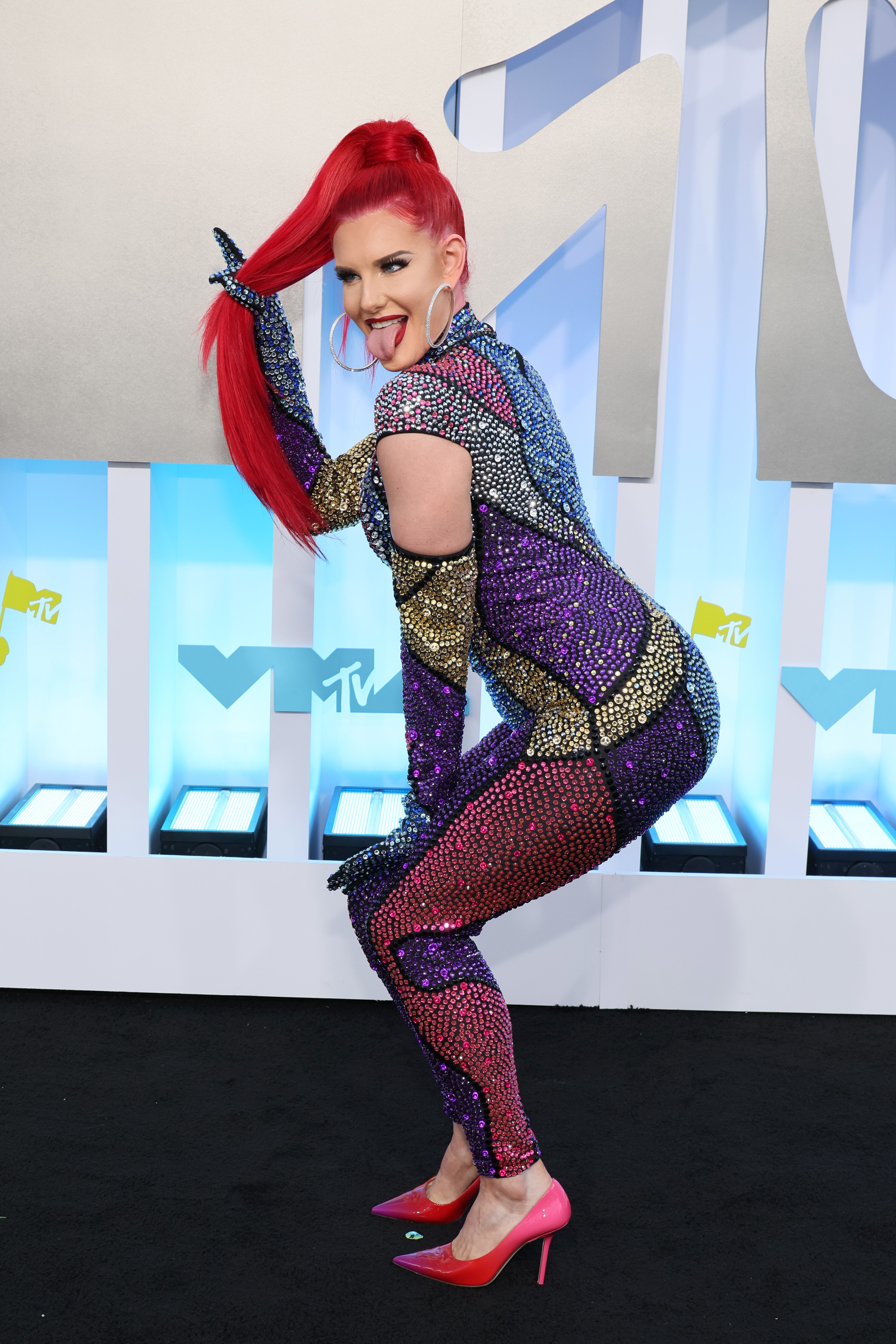 justina valentine poses on an arrivals carpet holding her bright red ponytail and sticking her tongue out
