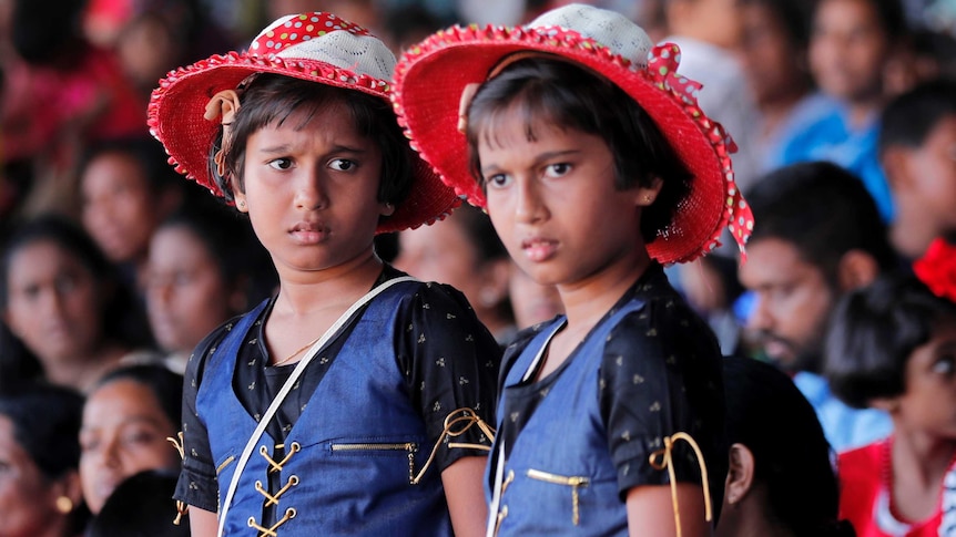 Two young twin girl wearing the same vest, dress and white and red hat standing among other twins.