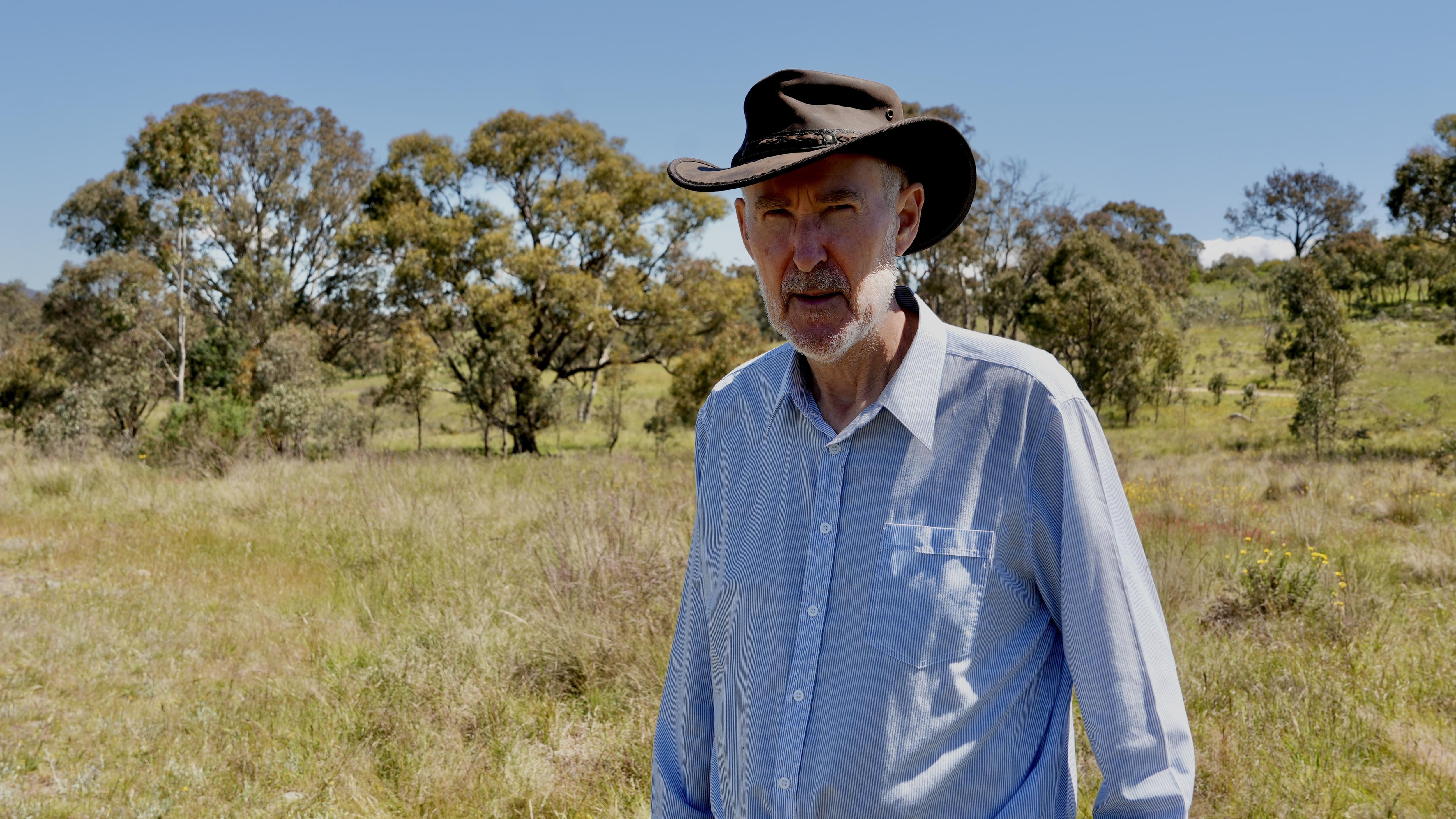 A man wearing an akubra standing on a bush property looking frustrated.