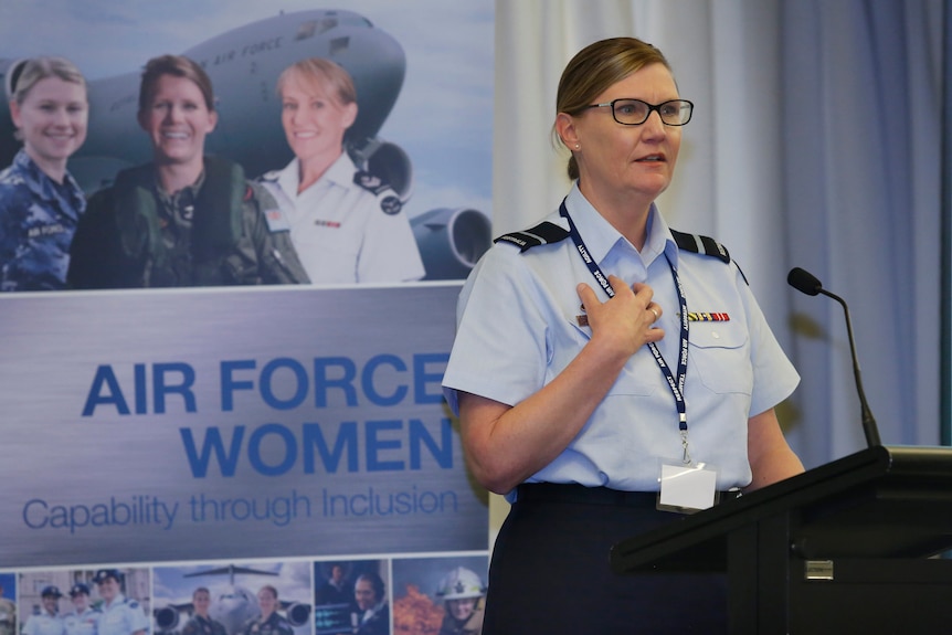 Kobieta w okularach ma na sobie jasnoniebieską koszulę munduru Sił Powietrznych i włosy upięte w kok, rozmawiając za wykładem