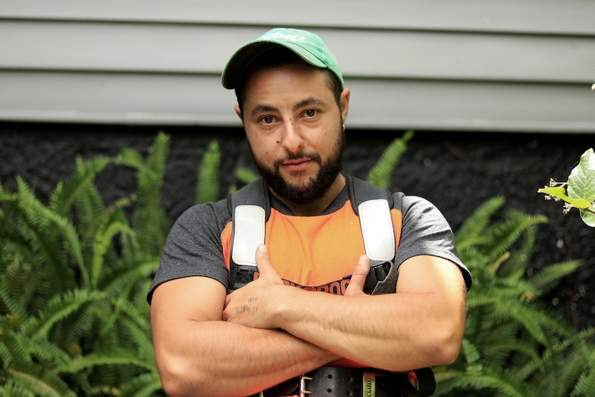 A young, bearded man stands outside with his arms folded wearing a green cap and orange and grey t-shirt.