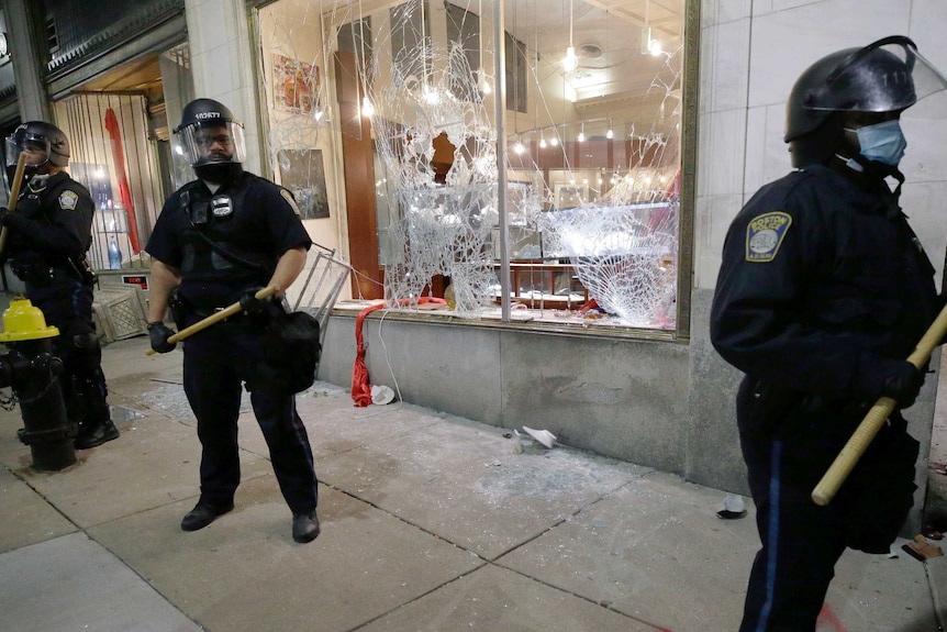 Police officers wearing helmets with face shields brandish wooden clubs as they guard a shop with a broken window.