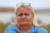 A woman with grey hair and a blue collared long sleeve shirt looks directly into the camera