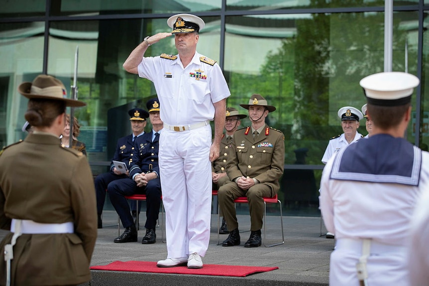 A high-ranking naval officer in white dress uniform salutes defence personell from a stage.
