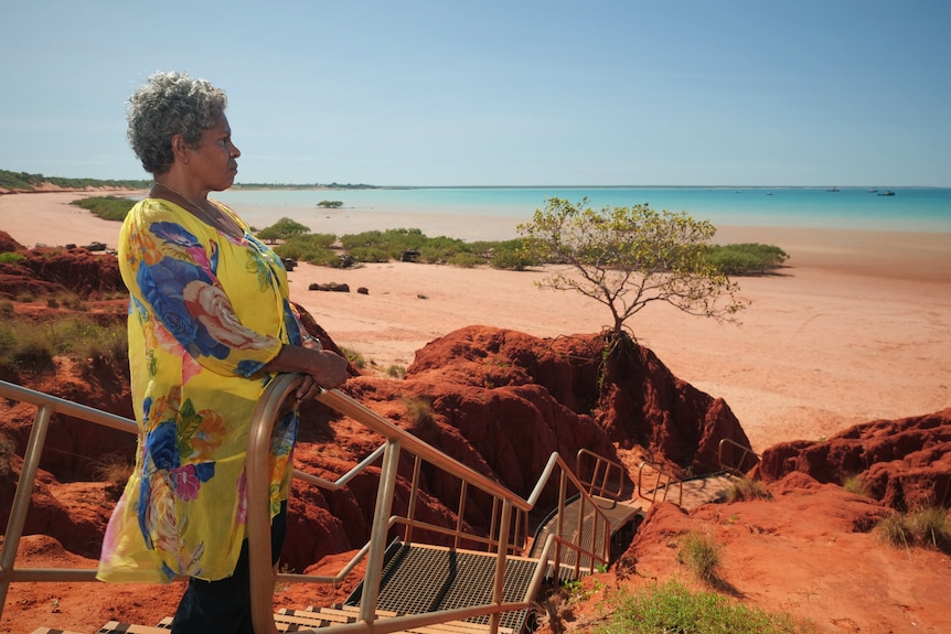 Vennessa Poelina stands among red rocks looking out towards the beach.