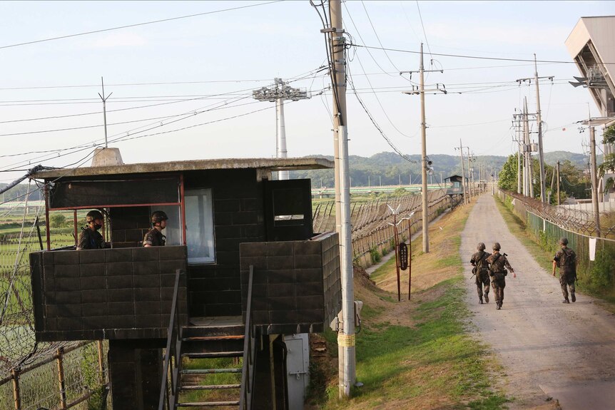 Two soldiers stand in a guard tower along a fortified border lined with high fences, while three soldiers patrol a road.