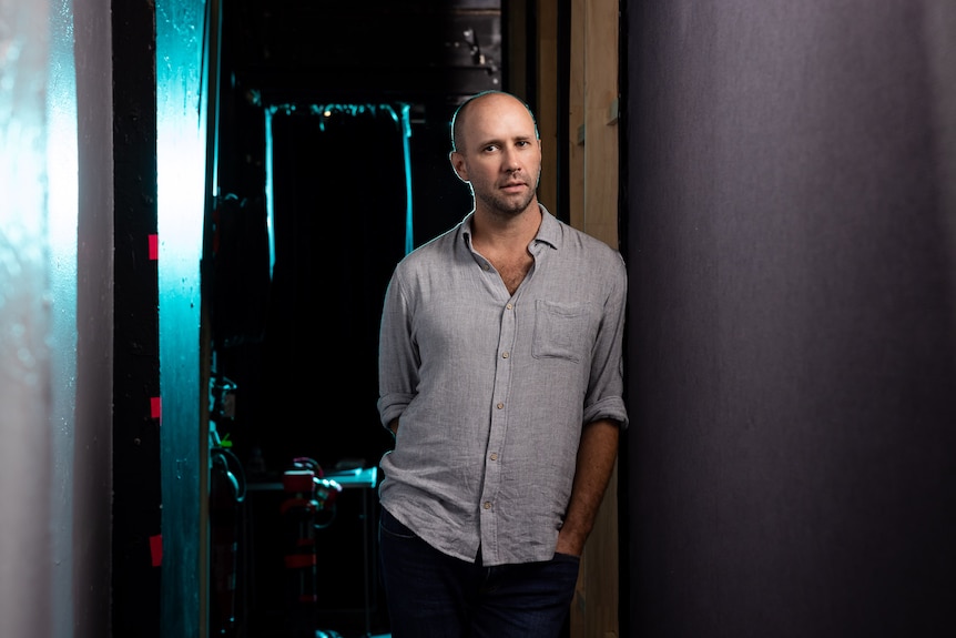 Bald man with light stubble and soft brown eyes looks pensively at the camera while leaning against a backstage wall.