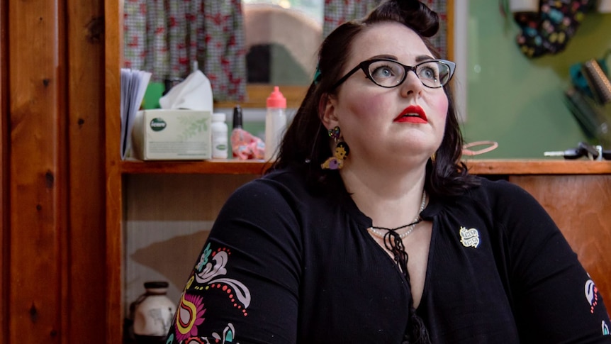 A woman with black hair, red lipstick and black glasses sits inside and looks upwards contemplatively.