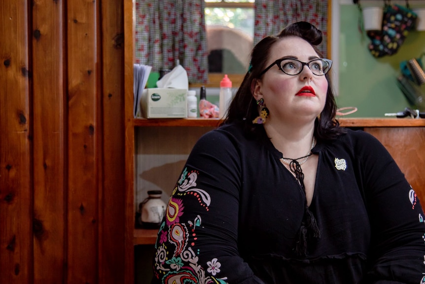 A woman with black hair, red lipstick and black glasses sits inside and looks upwards contemplatively.