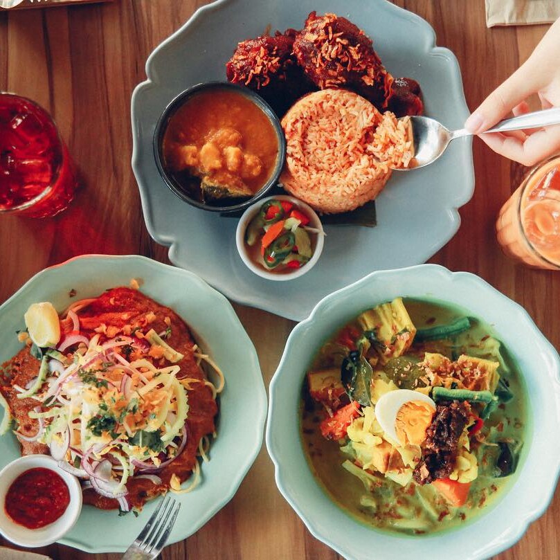 Overhead shot of three plates of Malay food and two drinks.