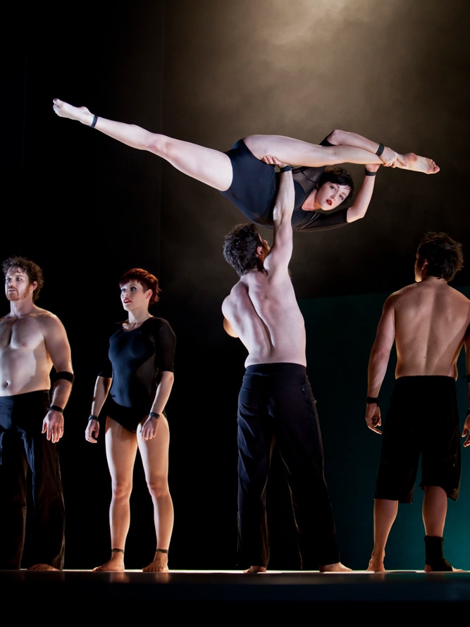 Five performers on stage, one of them (a man) holding another (a woman) aloft.