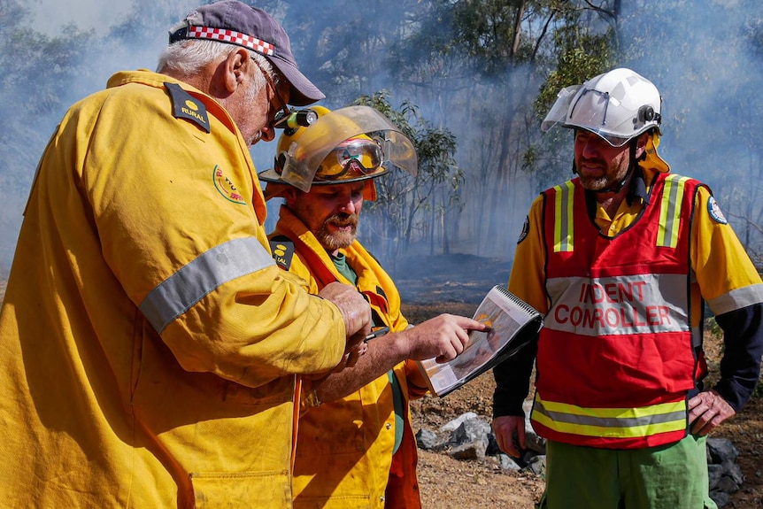 Three men in PPE gear look at a map while a fire burns behind them.