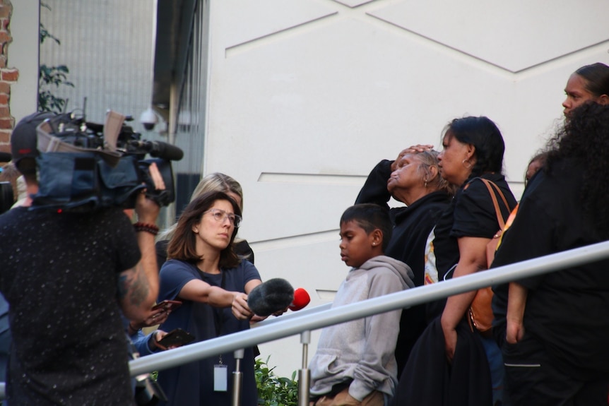 JC's foster mother puts her hand on her head, TV reporters interview the family.