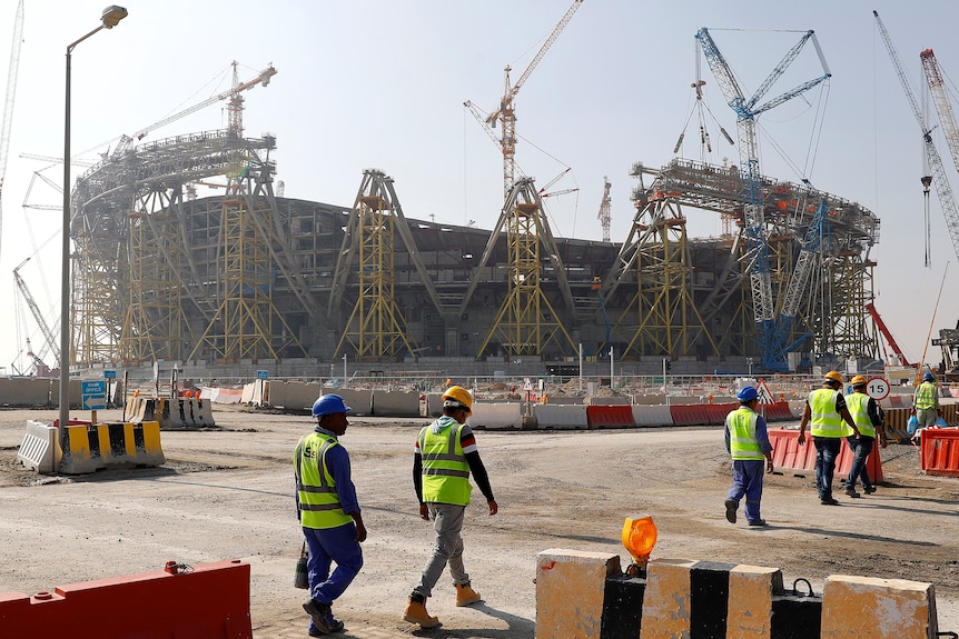 six workers in hi-vis yellow vests in foreground, a partially complete football stadium in background