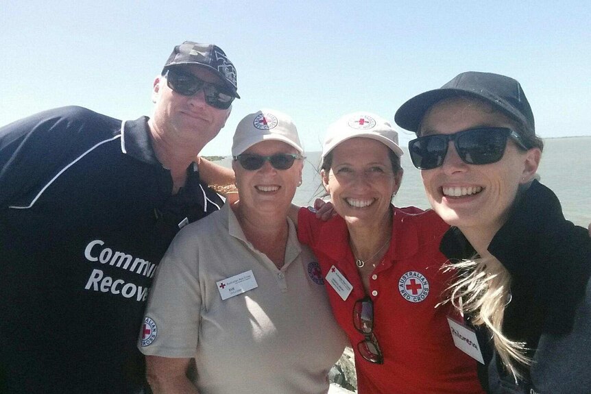 Three women and a man stand with their arms around each others shoulders. Two of the women wear Red Cross caps and T-shirts.