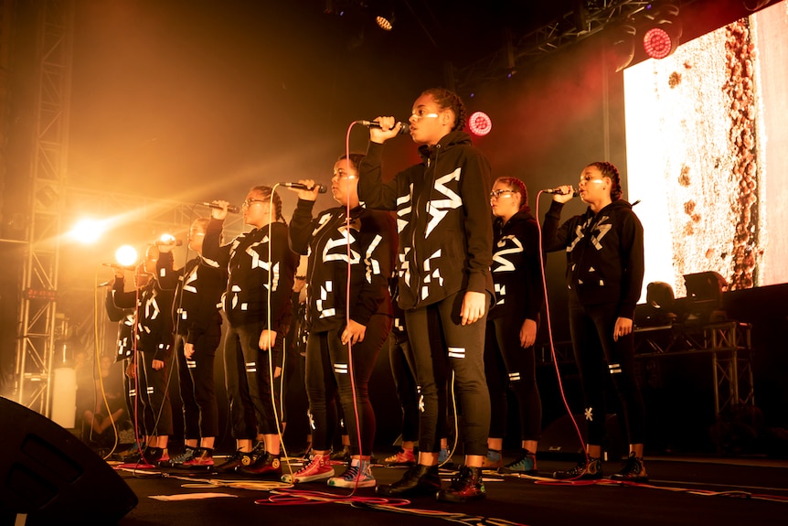 Two rows of teenage girls standing on orange-lit stage, wearing back tracksuit hoodies and with white ochre stripes under eyes.