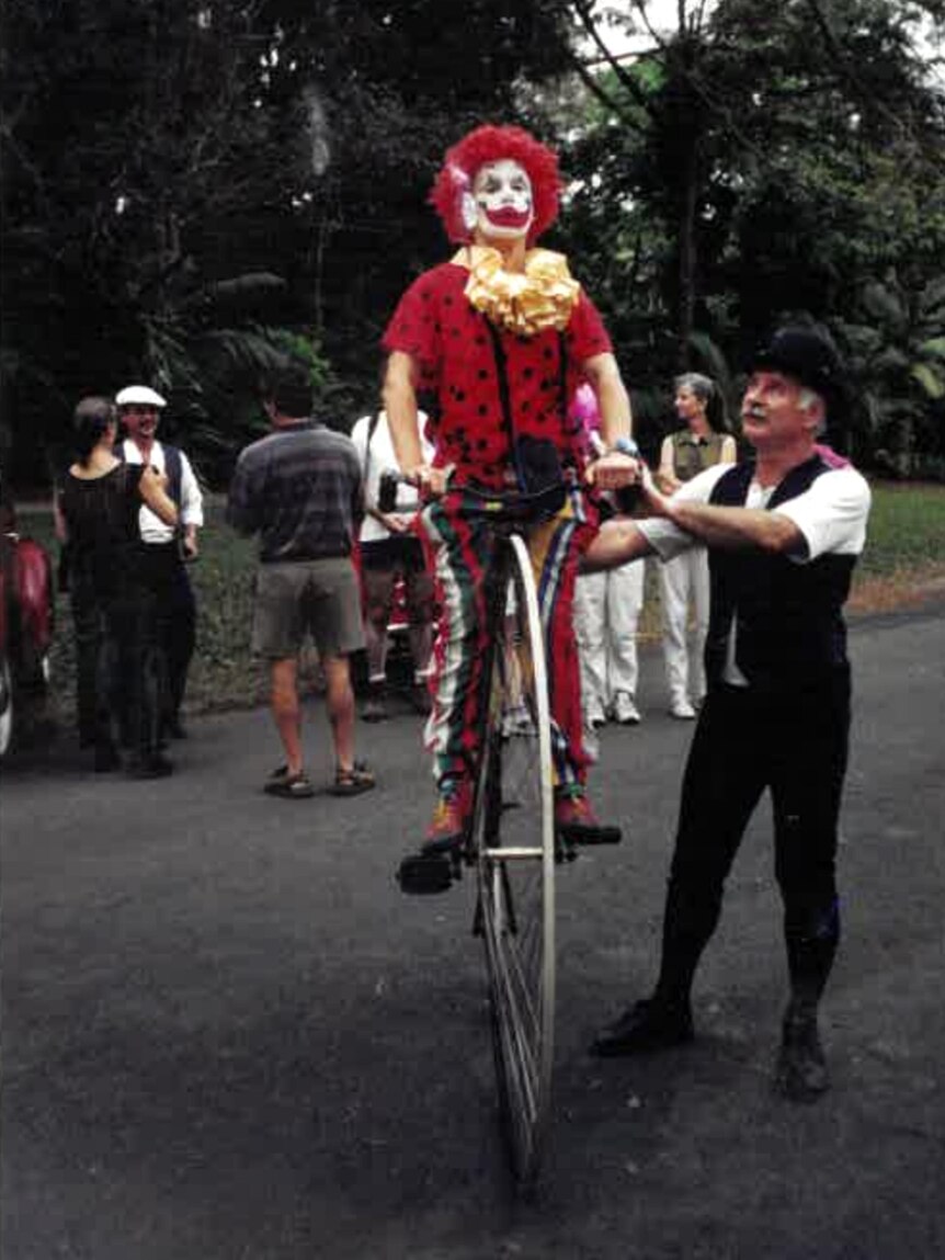 A clown sitting on a pennyfarthing, being supposed by a man in old fashioned dress standing next to her.