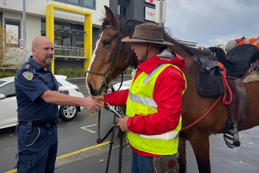 A paramedic in uniform shaking hands with a guy in an Akubra standing by his horse in a city setting.