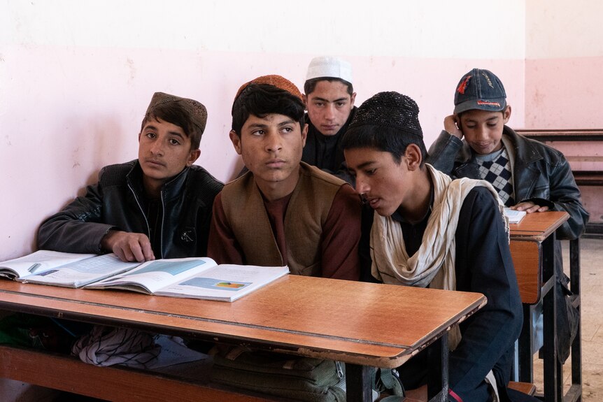 Boys crowd around a desk in a classroom and look at a book