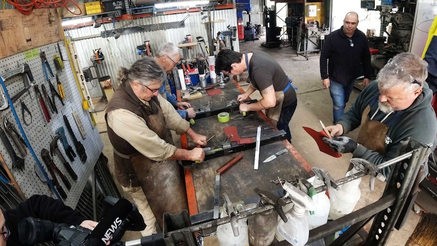 A group of men make knives around a workshop bench.