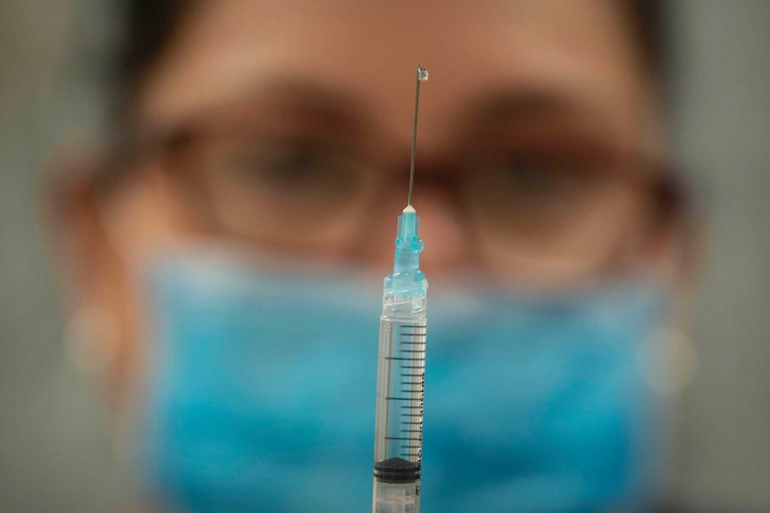 A person wearing a face mask and rubber gloves holds up a syringe