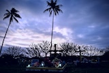 Crosses mark tombs at a cemetery, a grey cloudy sky and palm trees in the background. 