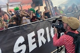 Cameraman filming crowd of people wearing green wigs and face paint at festival.