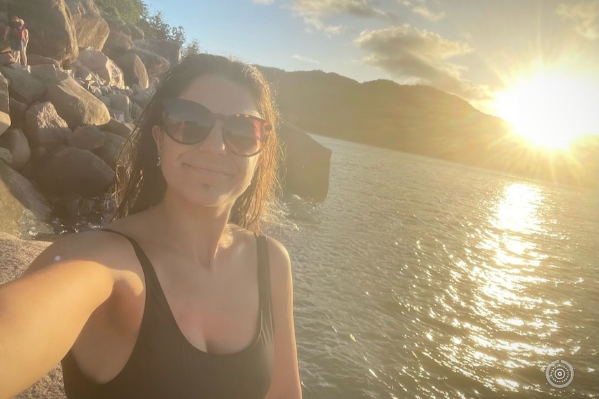 A woman in a black swimsuit and sunglasses on a beach with the sun setting in the background.