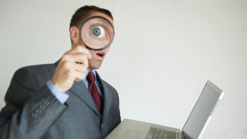 A man holding a magnifying glass in front of his eye while holding a laptop computer.