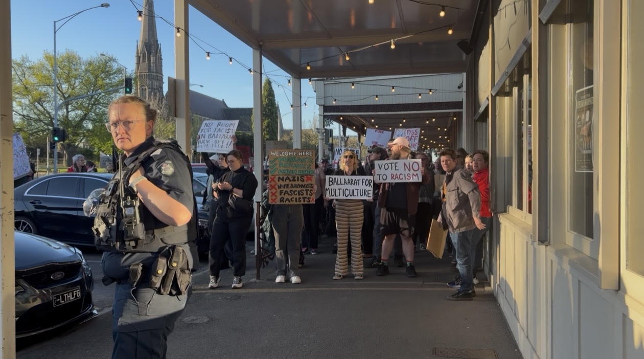 Protesters holding placards stand on a broad footpath in a country town.