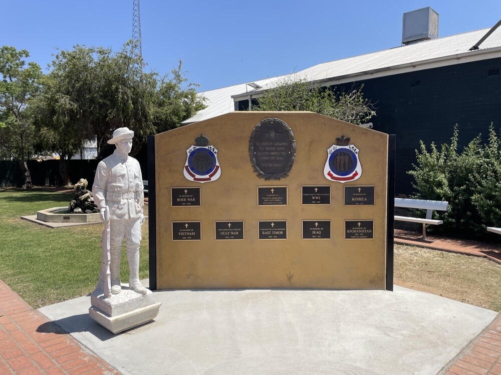 Uma estátua de pedra fica em um pedestal próximo a uma parede com placas comemorativas em homenagem aos veteranos de várias guerras.
