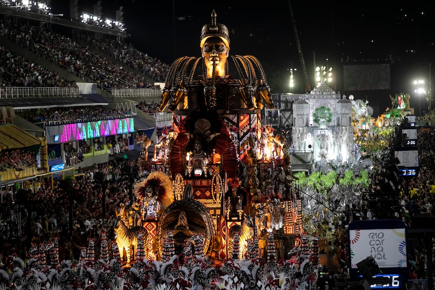 performers in colourful costumes dance on and around two large floats as they pass past stands fill with audience members