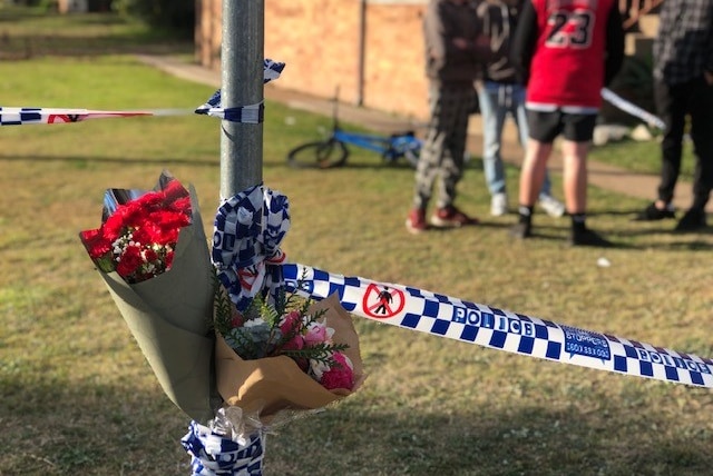 Bunches of flowers rest against a pole that has police tape tied around it. People are gathered in the background.