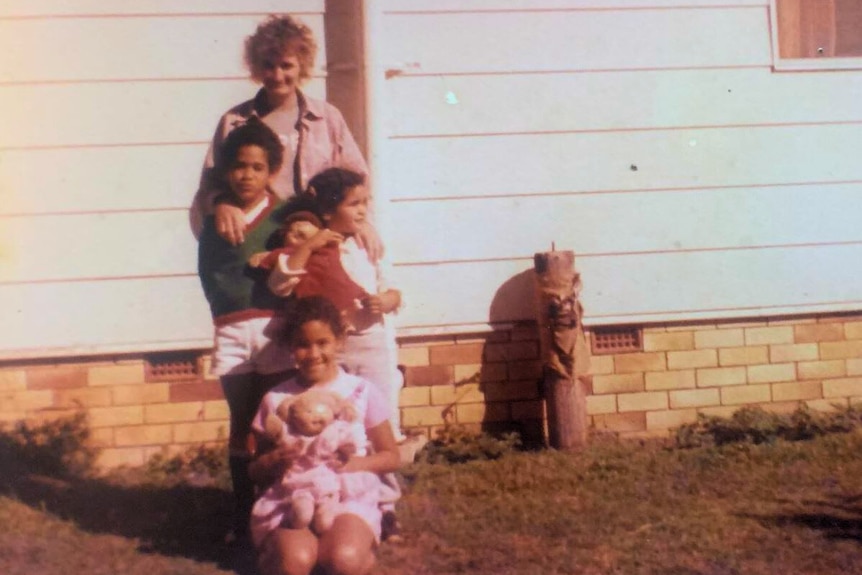 1980s photograph of a woman standing outside a home with a son and daughter and another daughter kneeling holding a doll