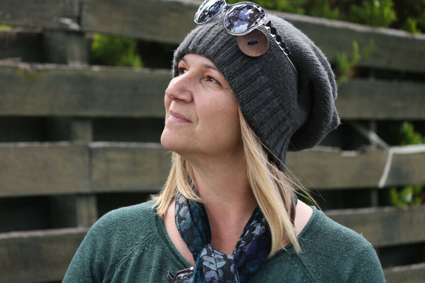 A woman with blonde hair wears a dark grey beanie with sunglasses on her head and looks off into the distance.