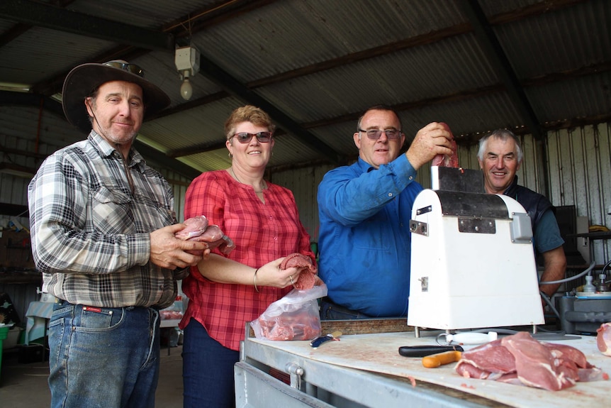 Four people stand in a row behind a ute, three men and one woman look at the camera. One feeds a steak through a meat tenderiser