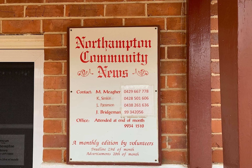 Sign on a red brick wall, in red writing 'Northampton Community News' with a list of names and phone numbers below.