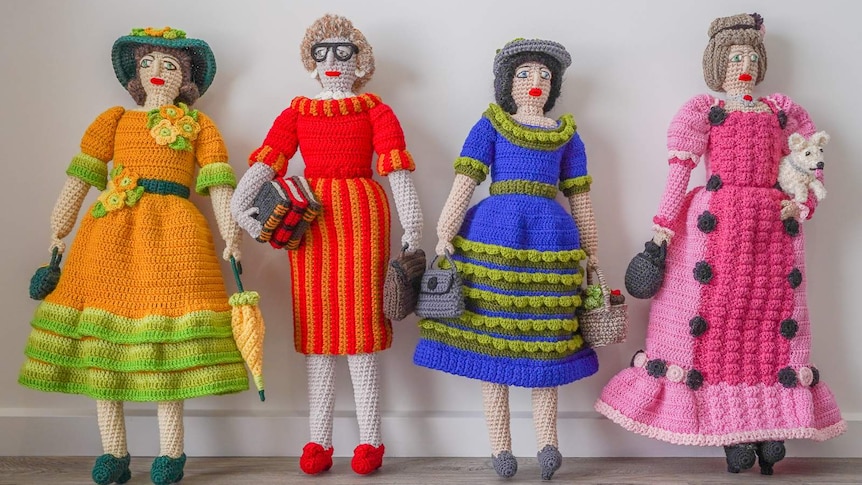 Four crochet ladies in colourful dresses and headwear stand against a white wall.