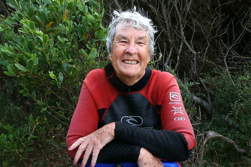 Portrait of Alison Parker, retired woman in her wetsuit leaning on a boogie board with tea trees behind