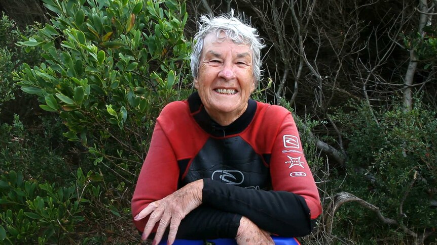 Portrait of Alison Parker, retired woman in her wetsuit leaning on a boogie board with tea trees behind